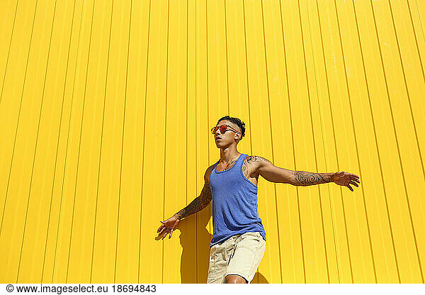 Young man with arms outstretched dancing in front of yellow wall on sunny day