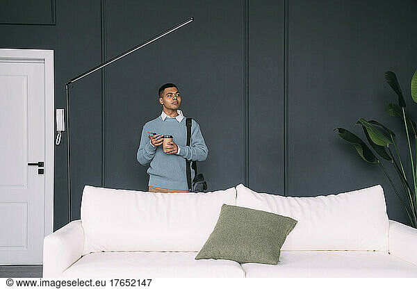 Young man with disposable coffee cup standing in living room