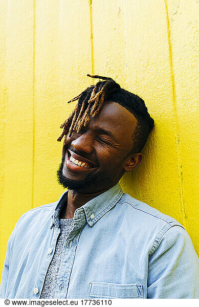 Young man with dreadlocks laughing in front of yellow wall