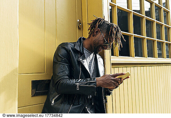 Young man with dreadlocks using smart phone by building