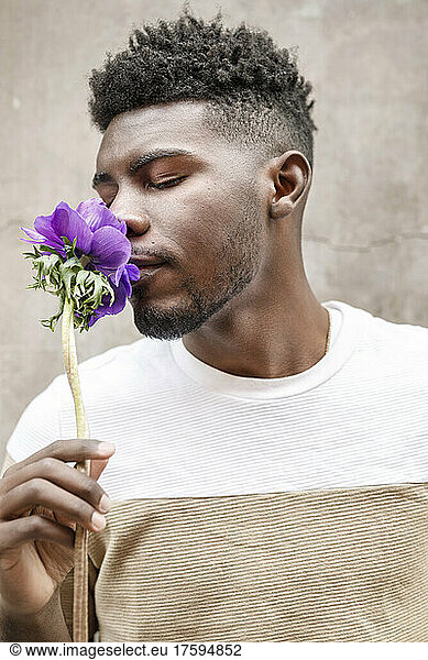 Young man with eyes closed smelling flower in front of wall