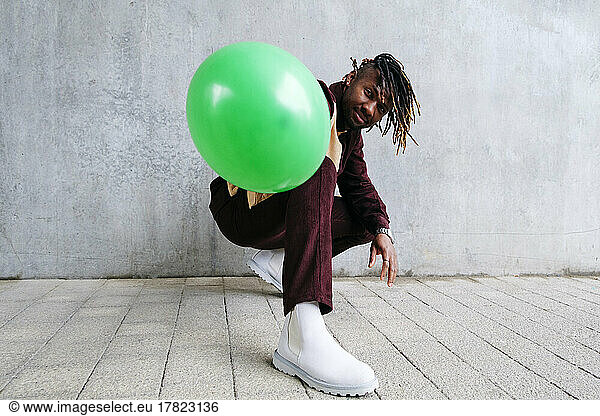 Young man with green balloon crouching in front of concrete wall