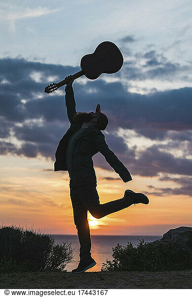 Young man with hand raised holding guitar during sunset