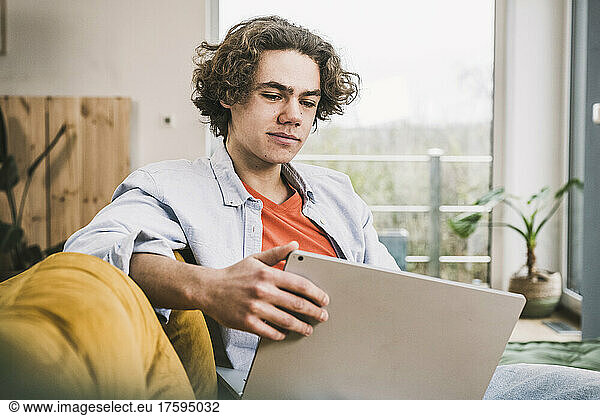 Young man with laptop sitting on sofa in living room at home