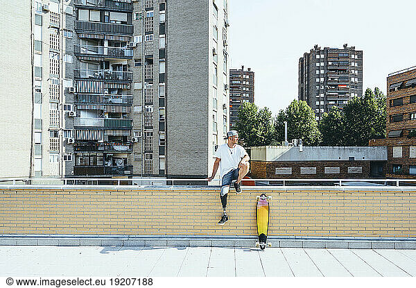 Young man with leg prosthesis sitting on railing in the city next to skateboard