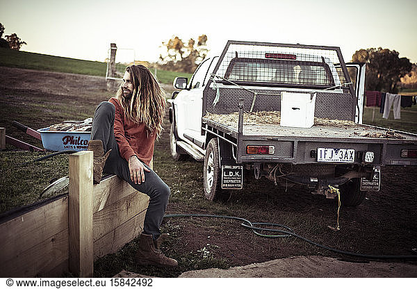 young man with long hair and beard sits on fence by farm truck