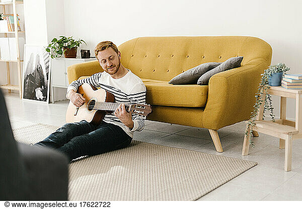 Young man with red hair practicing guitar sitting in front of sofa at home