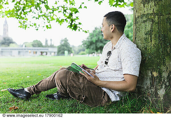 Young man with Sudoku book leaning on tree trunk at park