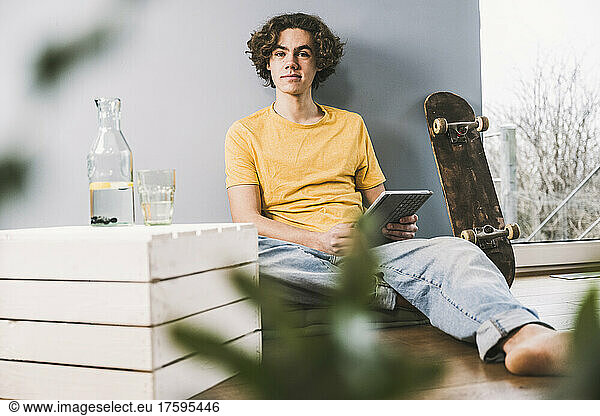 Young man with tablet PC sitting by skateboard at home