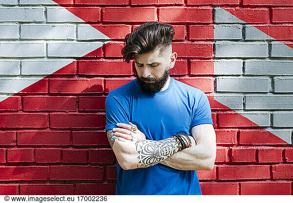 Young man with tattoo standing in front of red brick wall with arms crossed