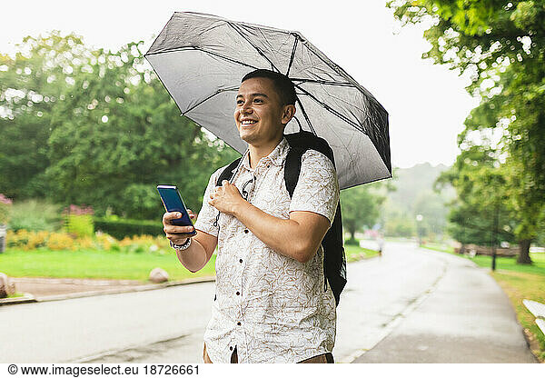 Young man with umbrella holding smart phone at park