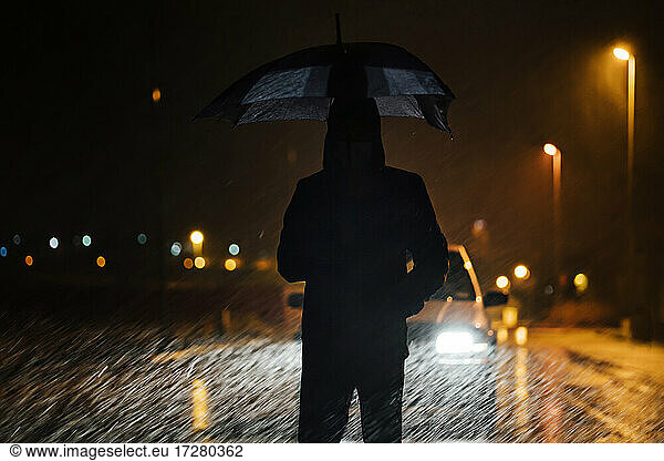Young man with umbrella standing in front of car dutimng a rainy night
