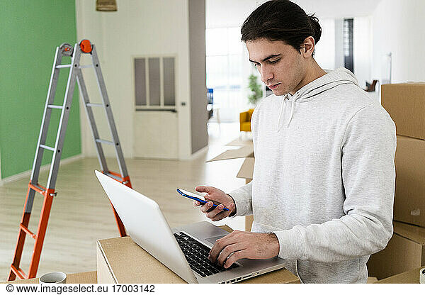 Young man working on laptop while using mobile phone while relocating in new house