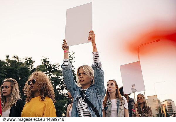 Young men and women protesting with posters while marching in city against sky