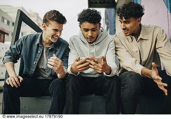 Young men looking at friend using smart phone while sitting outdoors