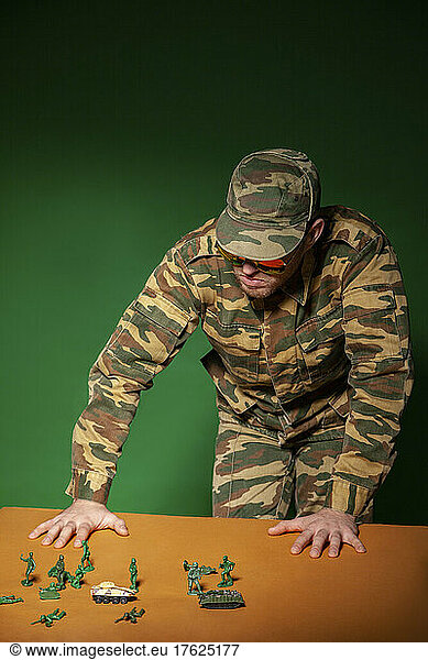 Young military soldier with army figurine toys standing by table against green background