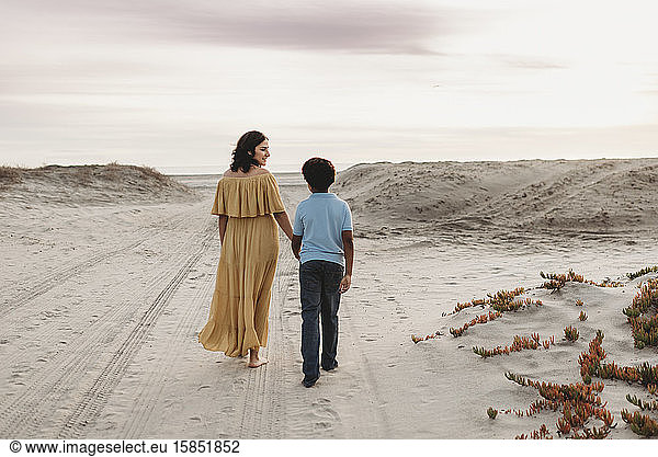 Young mother and school-aged son walking at beach against cloudy sky