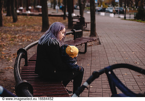 Young mother holding baby son while sitting on bench in park