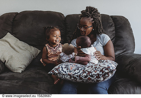 Young multiracial girl smiling at infant sibling on couch with mom