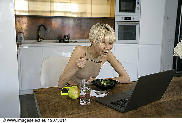 Young nutritionist eating salad and using laptop at dining table in kitchen