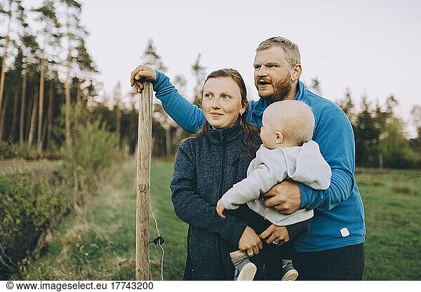 Young parents with toddler son standing by fence in farm during sunset
