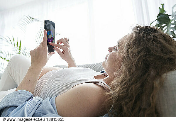Young pregnant woman using smart phone lying on sofa at home