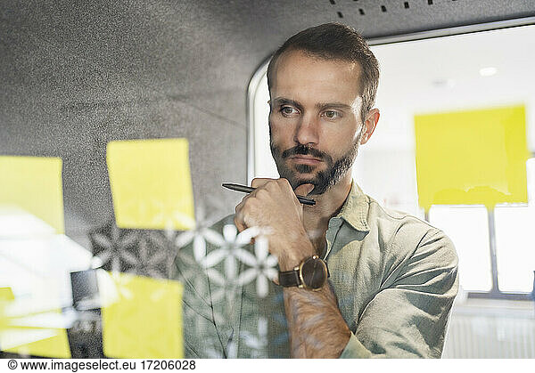 Young professional contemplating over adhesive note on telephone booth glass at office