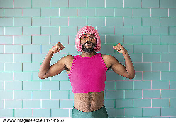 Young queer man flexing muscles standing in front of wall