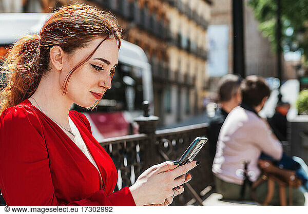 Young red-haired girl dressed in a red blouse using her mobile phone in a city square.