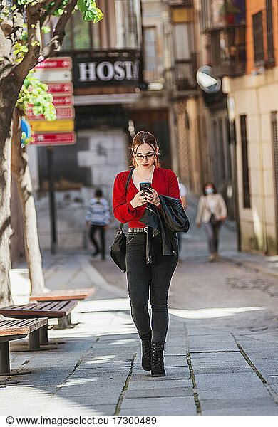 Young red-haired girl with glasses dressed in a red blouse and jeans walking down a city street while using her mobile phone