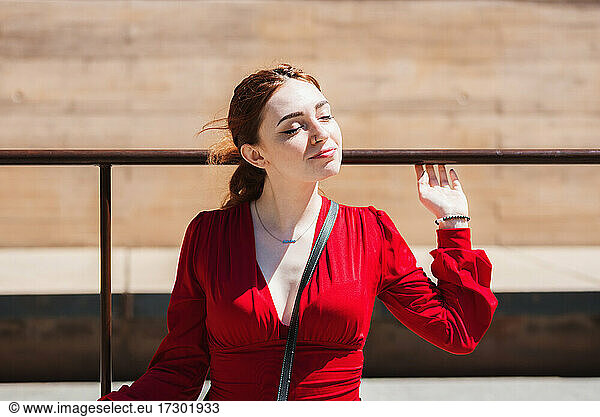 Young red-haired woman enjoying the sun in the city. Dressed in a red blouse.