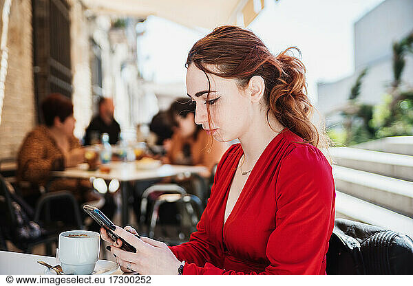 Young red-haired woman in a red blouse drinking coffee on a terrace while using her mobile phone.
