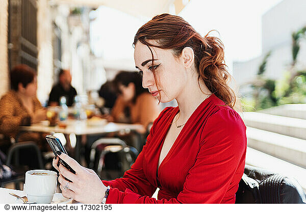 Young red-haired woman in a red blouse drinking coffee on a terrace while using her mobile phone.