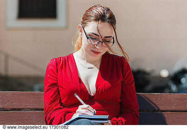 Young red-haired woman in a red blouse sitting on a bench while writing something in her notebook.