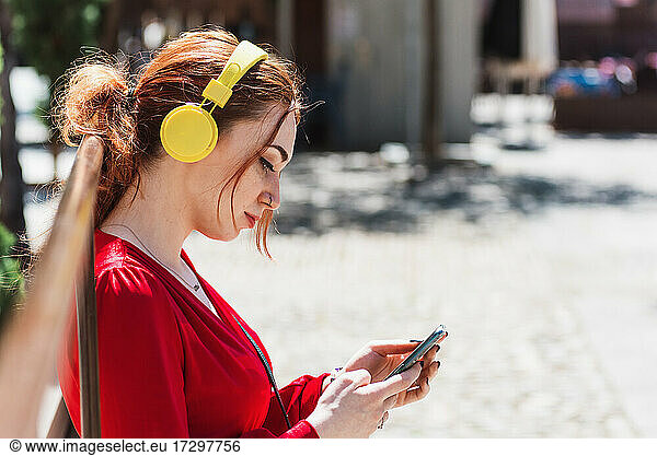 Young red-haired woman listening to music with her cell phone and yellow headphones in an urban space. Dressed in a red blouse.