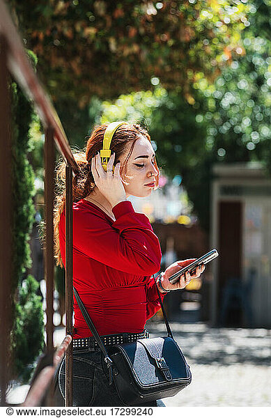 Young red-haired woman relaxing while listening to music with her cell phone and yellow headphones in an urban space. Dressed in a red blouse.
