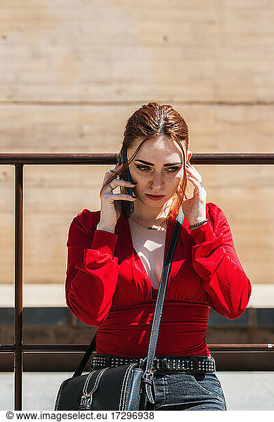 Young red-haired woman talking on mobile phone in an urban space. Dressed in a red blouse.