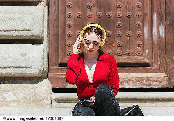 Young red-haired woman with a red blouse sitting while listening to music with her cell phone and yellow headphones. Old wooden door in the background.