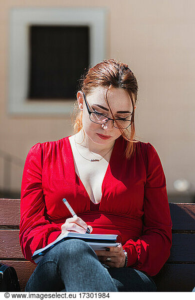 Young red-haired woman with eyeglasses and dressed in a red blouse sitting on a bench while writing something in her notebook.