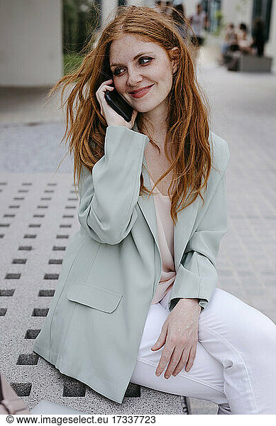 Young redhead businesswoman talking on smart phone while sitting on bench