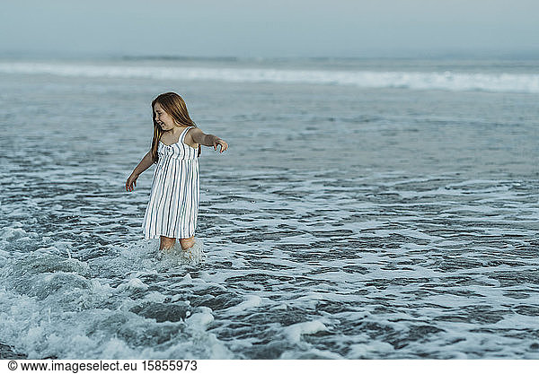 Young redhead girl in dress playing in ocean waves at dusk