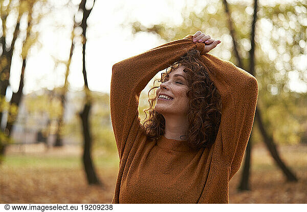 Young redhead woman with eyes closed and arms raised at autumn park