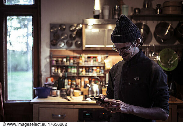 Young smiling man in glasses an beanie checks mobile phone in kitchen