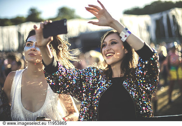 Young smiling woman at a summer music festival wearing multi-coloured sequinned jacket  taking picture with smartphone.