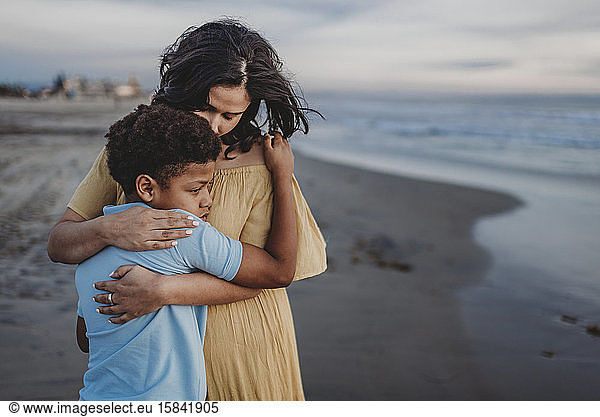 Young son and mother embracing at beach during sunset with cloudy sky