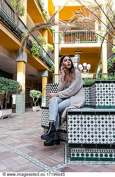 Young Spanish brunette woman with shopping bags sitting on a bench and talking on her cell phone.