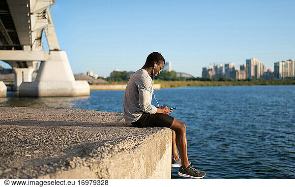 Young sportsman sitting with smartphone by river