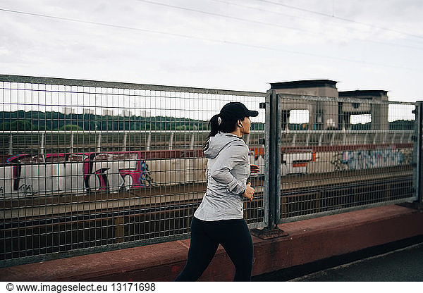 Young sportswoman jogging by fence on bridge in city
