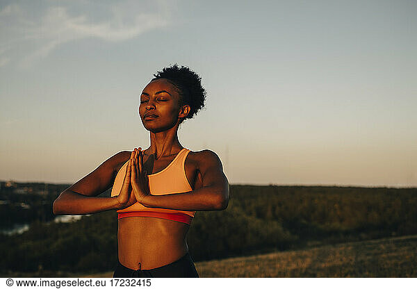 Young sportswoman meditating against sky during sunset