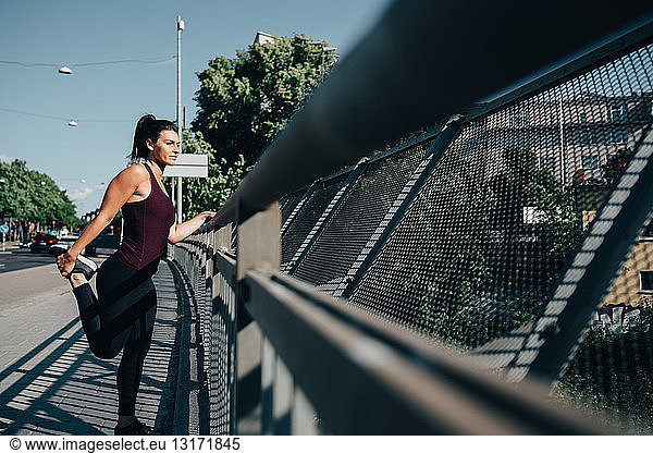 Young sportswoman stretching leg by railing on bridge in city